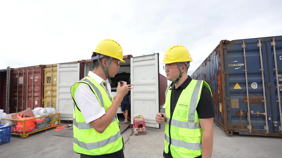 Two people wearing helmets and safety vests talking at a container yard